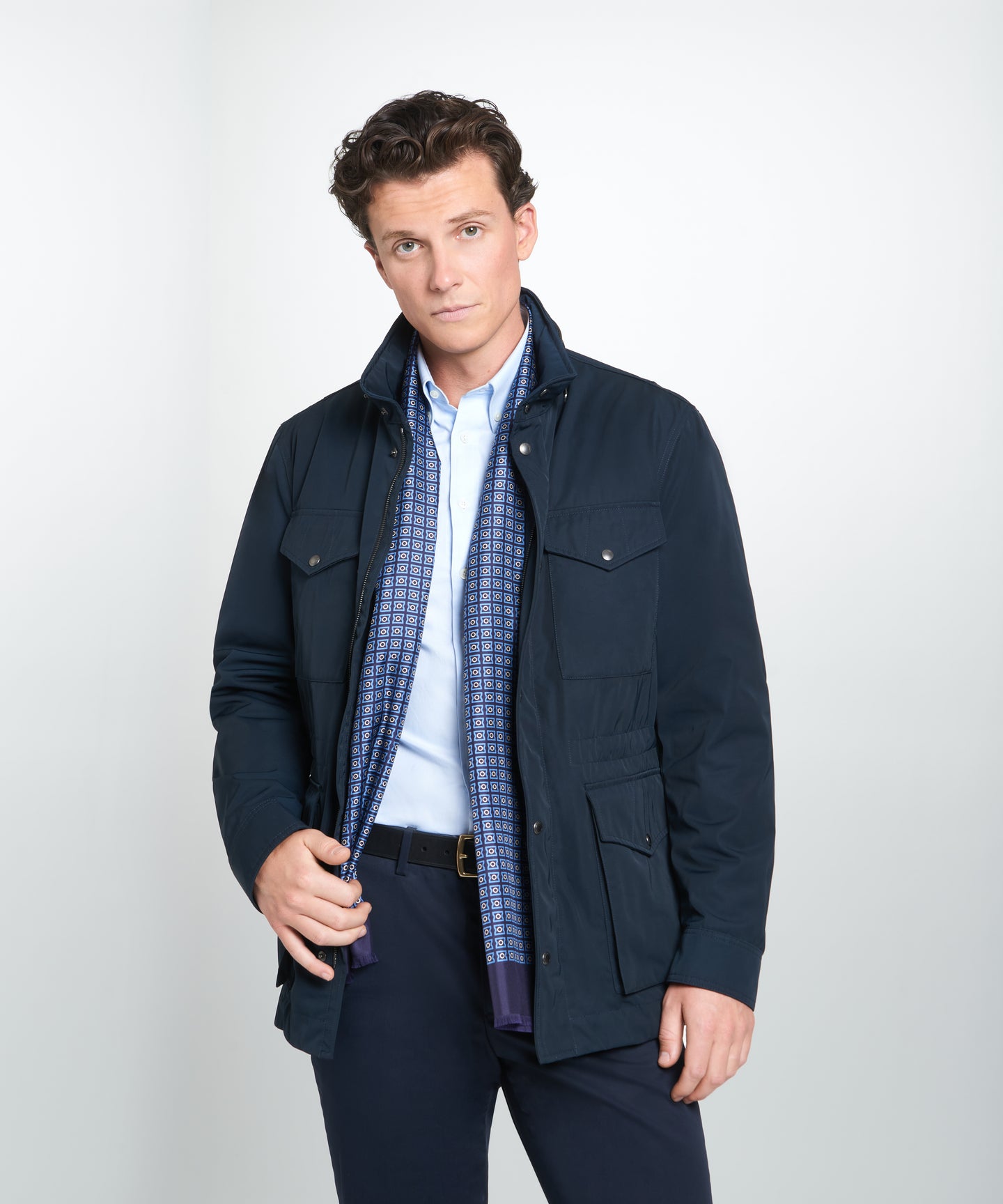 Man wearing a navy jacket with a checkered silk scarf and blue button shirt, on a cool grey background.